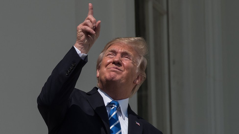 Trump señalando al cielo y mirando sin lentes el eclipse.