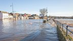 A street overrun with floodwater. The sky above is a pale blue