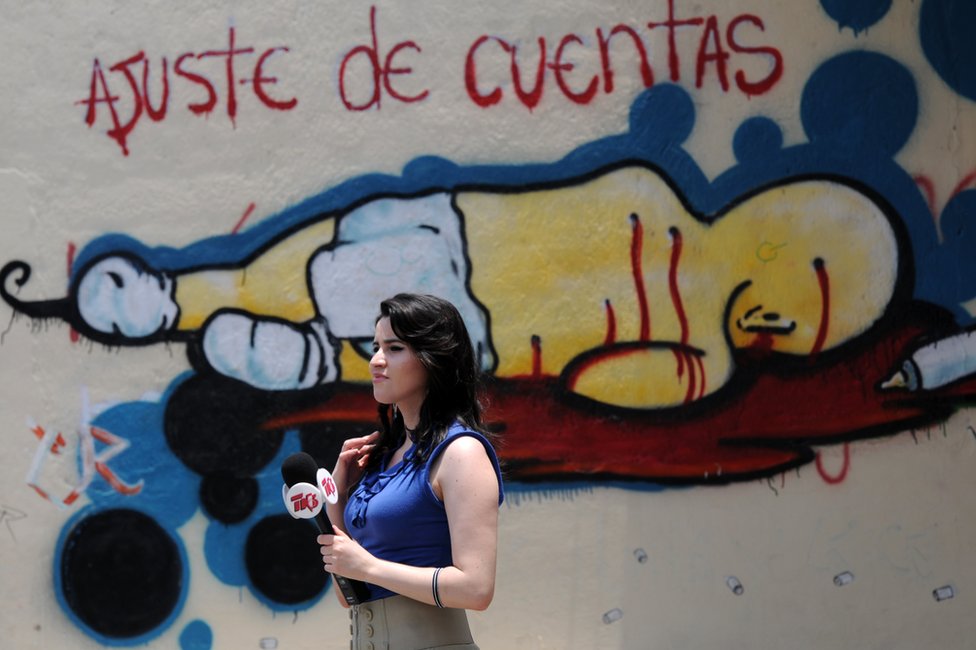 Periodista frente a un grafiti en el que se lee 