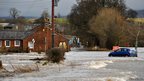 Flooding by houses. A "road closed" sign can be seen beside a blue car that sits in low floodwaters