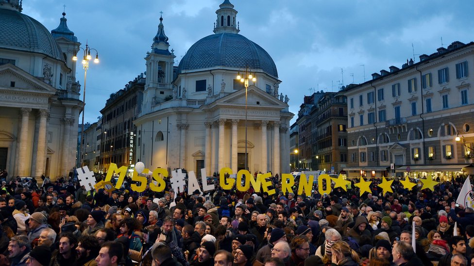 A large crowd gathers in a city square. Among them are individual letters arranged to say : "M5S al governo *****"