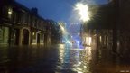A flooded street at night. Christmas lights can be seen strewn across the tops of the buildings