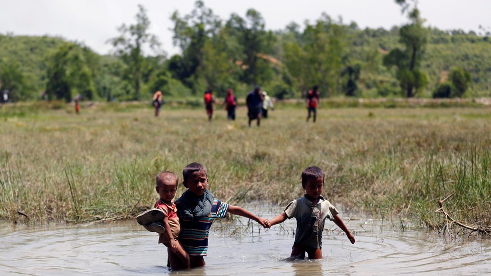 طفلان من الروهينغا يعبران النهر وهما ممسكان بإيديهما وأحدهما يحمل طفلاً صغيرا على ذراعه الآخر