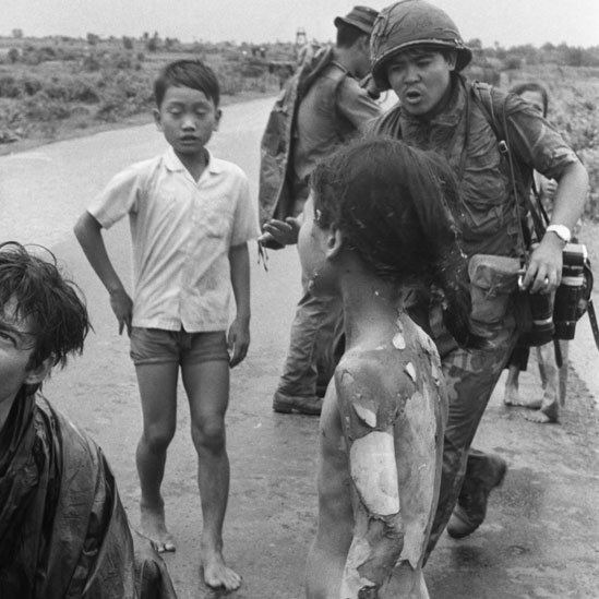 Niños tras el ataque de napalm en 1972 sobre la aldea de Trang Bang. Se ve a Nick Ut con sus cámaras colgando y a Kim desnuda de espalda con la piel que cae como en gajos