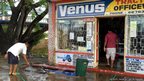 A man stands outside a shop, washing water out of the road.