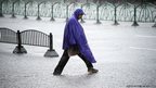 A man in a purple rainproof poncho crosses the road, under torrential rain. 