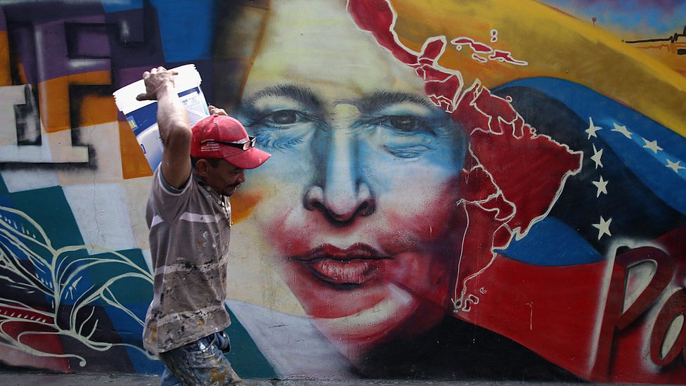 Hombre caminando en Caracas frente a un mural de Hugo Chávez.