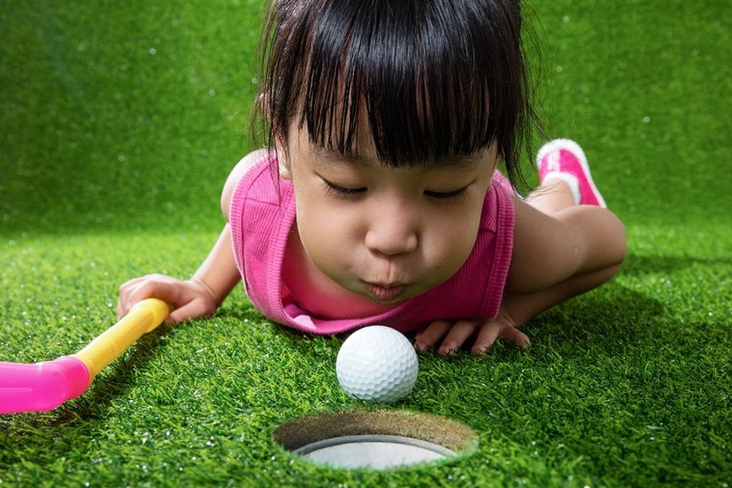 A little girl, lying on a mini-golf grass floor, blowing the ball into a hole