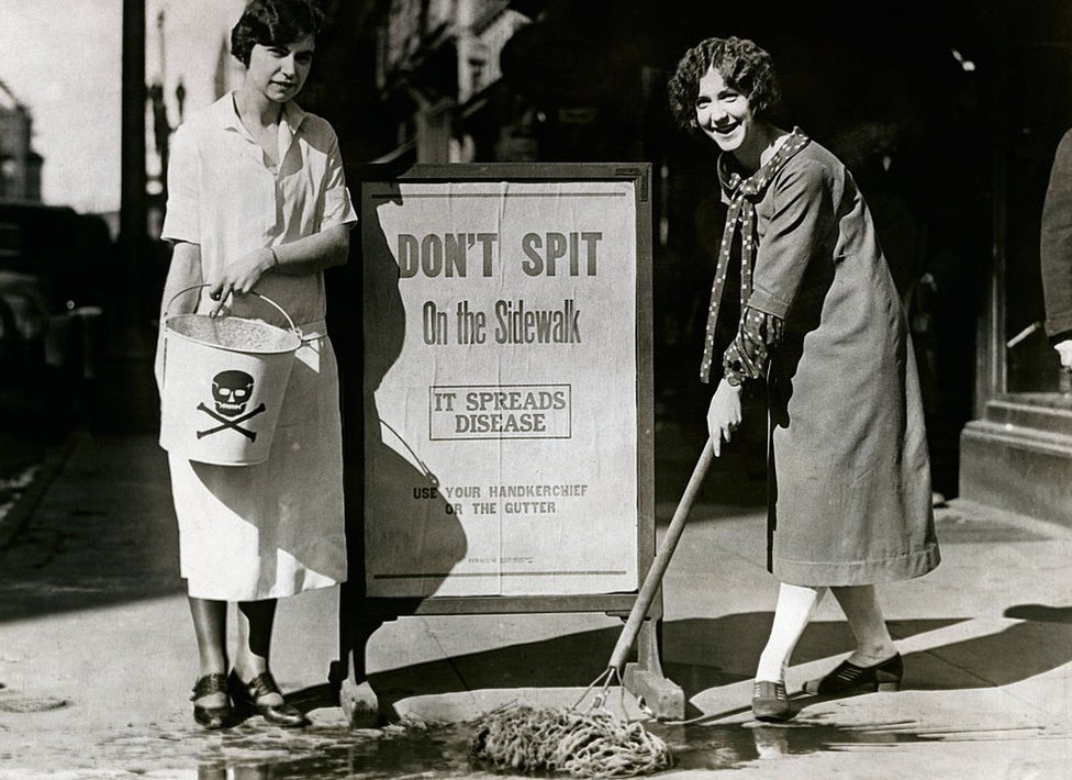 Em foto em preto e branco, duas mulheres ao lado de um cartaz onde se lê:''não cuspa na calçada, isso espalha doenças'