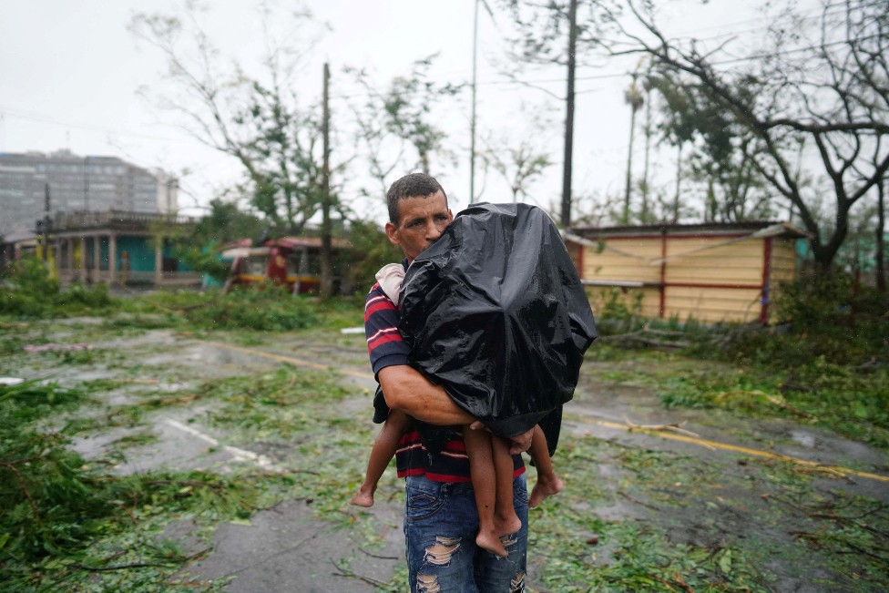 Un hombre carga a su hijo, al que lleva tapado con un impermeable negro y todo alrededor lleno de ramas rotas y destrozos.