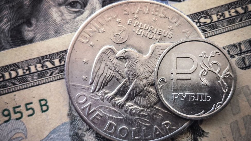 A Russian rouble coin is pictured with US dollar bills and a one dollar coin in Moscow, on March 15, 2022
