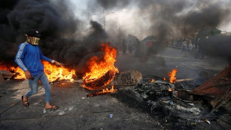 Демонстрант в маске идет по горящим шинам в Басре, южный Ирак
