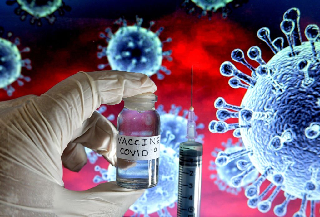 A syringe and a gloved hand holding a vile labelled "VACCINE COVID-19" with a background graphic illustration of a virus.
