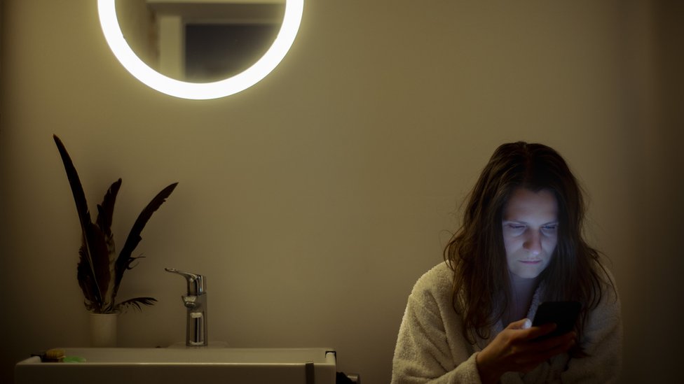 Una mujer revisa su teléfono dentro de un oscuro cuarto de baño
