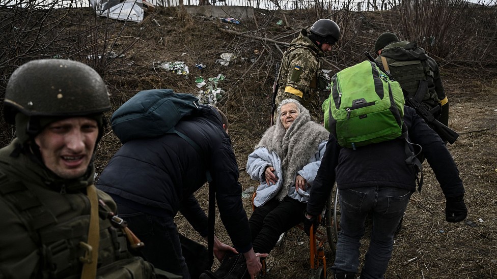 Una mujer de avanzada edad en silla de ruedas es cargada por soldados ucranianos