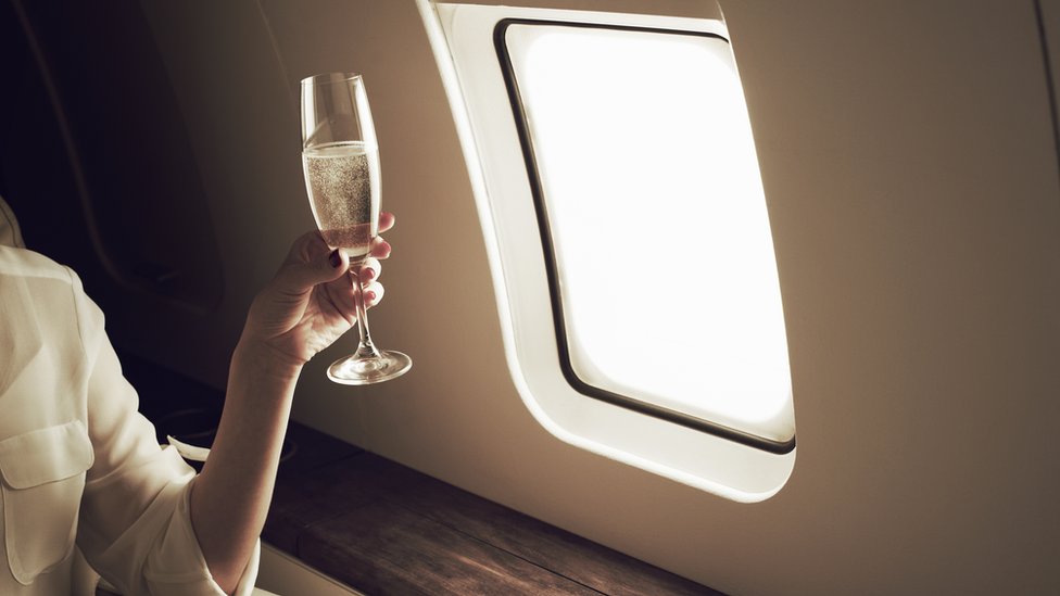 Mulher com um champanhe na mão, ao lado de uma janela de avião