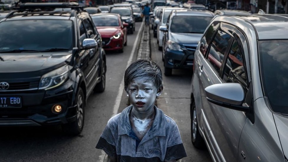 A boy whose skin appears metallic standing next to cars in traffic