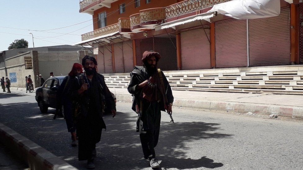 Três combatentes do Talebã caminham na rua, um deles com arma visível