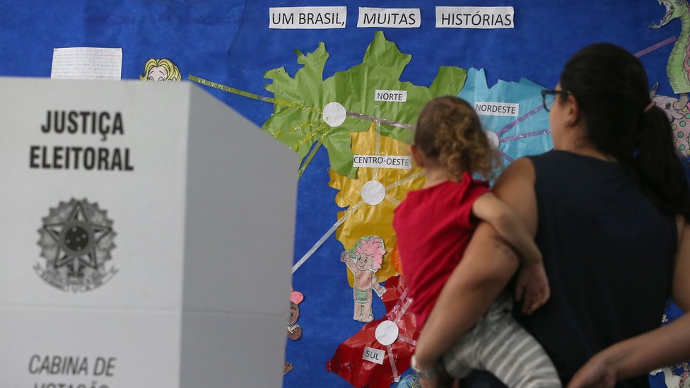 Eleitora com a filha no colo, ao lado de uma sessão de votação, e em frente a um mapa do Brasil com o nome das 5 regiões