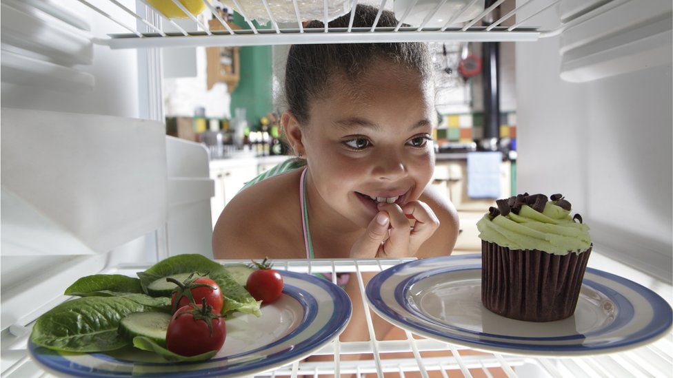 Menina olhando dentro da geladeira, onde está um prato de salada e um prato com cupcake