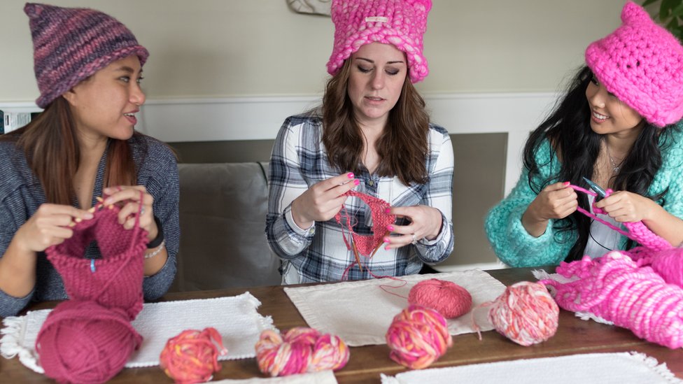 Видно, как три женщины в шляпах разговаривают друг с другом, вяжут еще больше за столом, уставленным клубками розовой пряжи