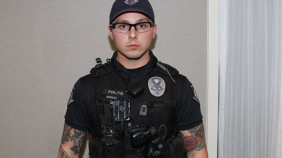 Филип Брейлсфорд, одетый в полицейское снаряжение, в кепке и очках. Его руки сильно татуированы, и он чисто выбрит