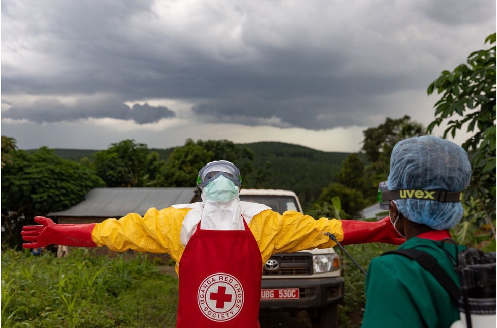 عامل في الصليب الأحمر يرتدي ملابس واقية ويقف وأذرعه ممدودة تحت سماء ملبدة