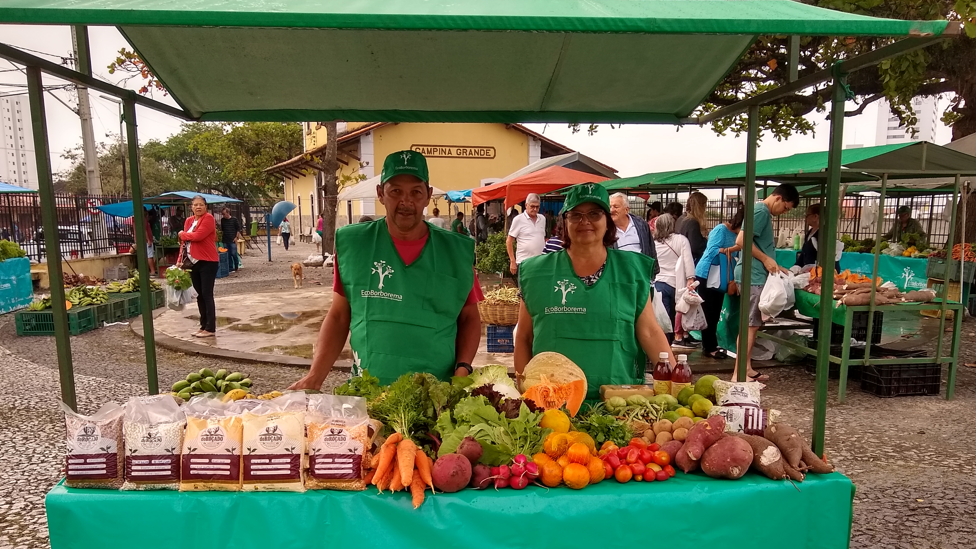 Duas pessoas atrás de barraca com frutas e legumes