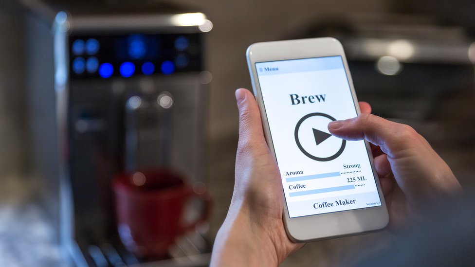 Ahora hasta las cafeteras están conectadas a internet (en la pantalla del celular se lee la indicación "hacer café"). Persona manejando a través de un celular una cafetera inteligente.