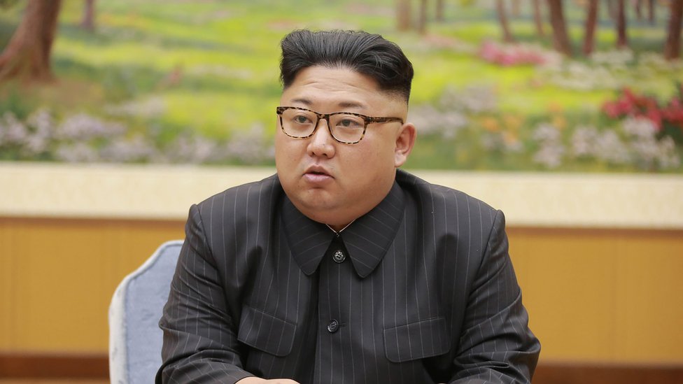 Ким Чен Ын сидит в коричневых очках черепаховой расцветки и темно-сером костюме в тонкую полоску