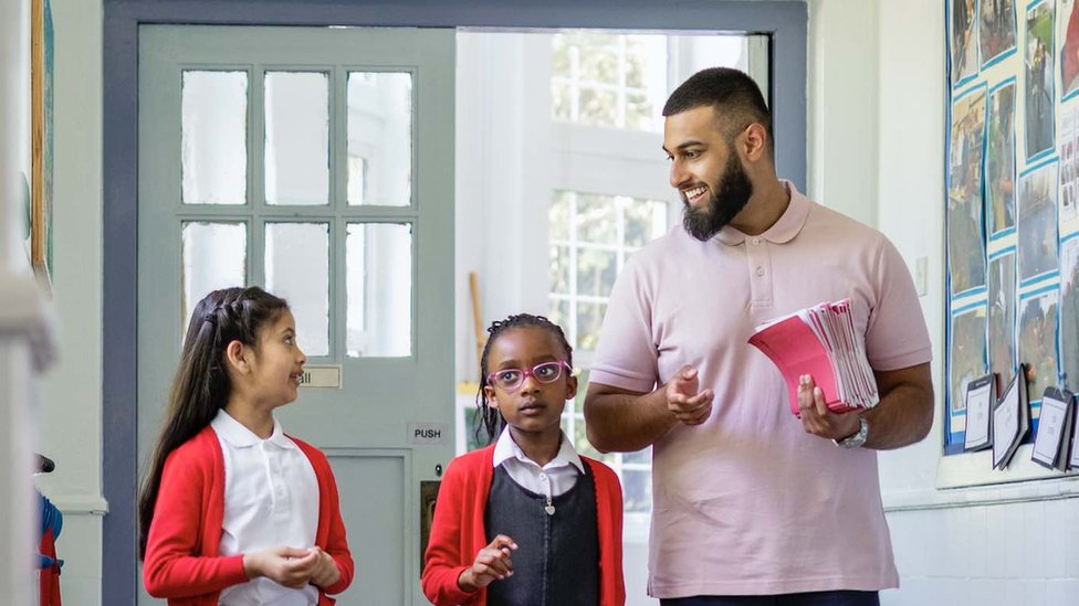 Two schoolgirls walk down a corridor with a teacher