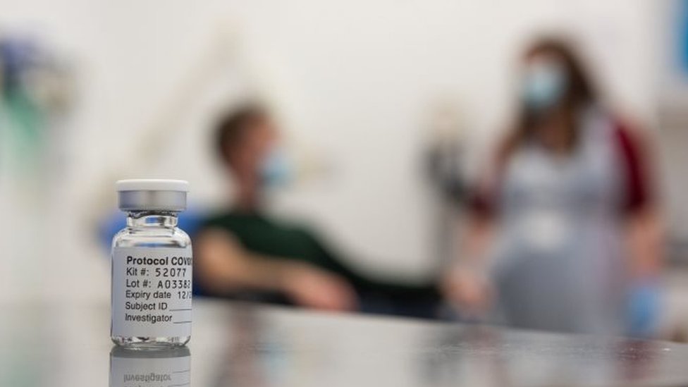 A test volunteer and medical worker behind a vial of the Oxford-AstraZeneca vaccine