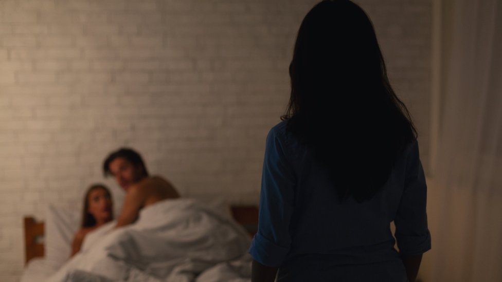 A woman looks at a man and a woman in bed