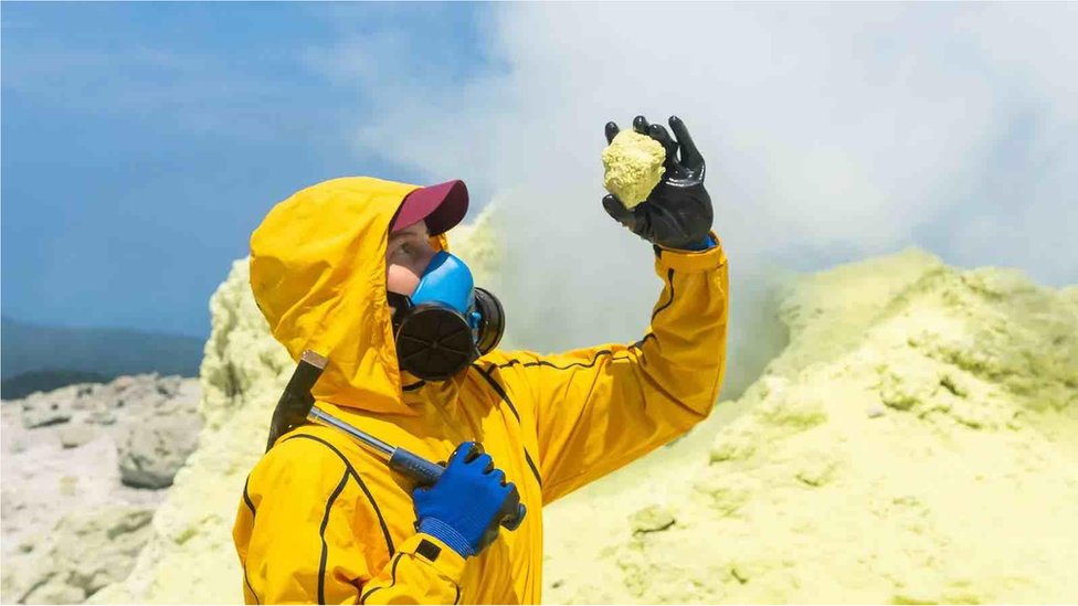 Woman volcanologist on the background of a smoking fumarole examines a sample of a sulfur mineral