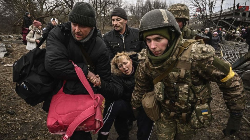 Uma mulher visivelmente exausta é assistida ao atravessar uma ponte destruída na cidade de Irpin, a noroeste de Kiev