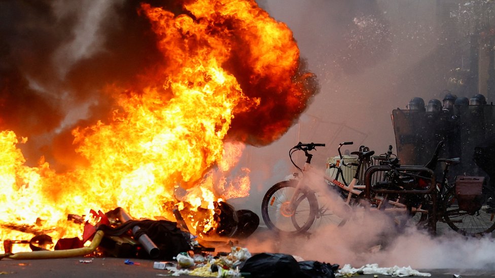 A large fire in the street in central Paris, with riot officers standing in a formation nearby