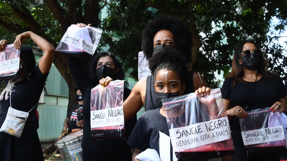 Mulheres seguram sacos plásticos com líquido vermelho e a frase 