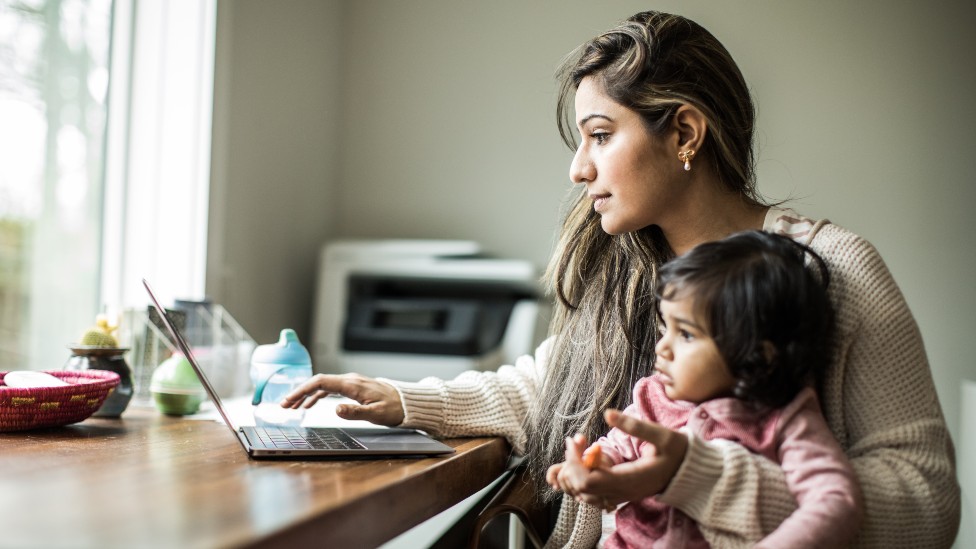 A mother holding her child at a computer while working from home