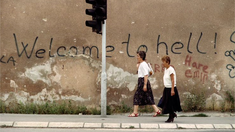 Two women walk in Sarajevo with a "Welcome to Hell" graffiti message in the background in 1994