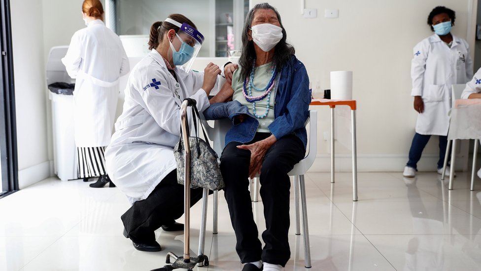 Mulher idosa de cbaço preto e cumprido, com colar de contas e olhos puxados, recebe vacina dada por uma profissional de saúde que usa uniforme escrito SUS