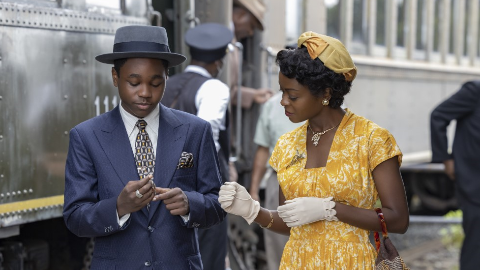 Fotografia colorida mostra um jovem negro de terno e chapéu e uma mulher negra de vestido amarelo, luvas e chapéu em uma estação de trem.