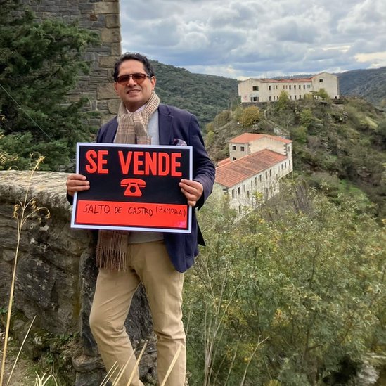 Ronnie Rodríguez con un cartel de "Se vende" y el pueblo de fondo
