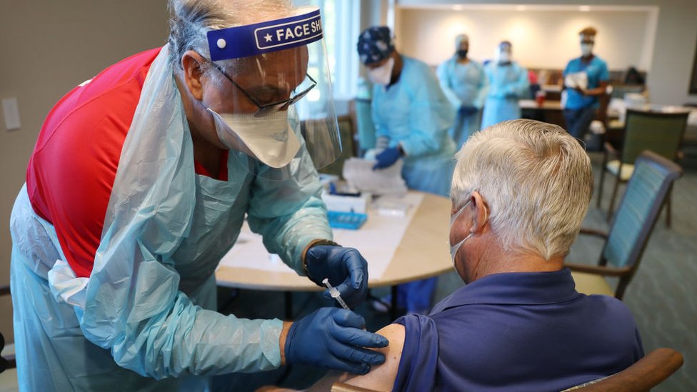 Enfermeiro aplica vacina em idoso, que aparece de costas dentro de sala