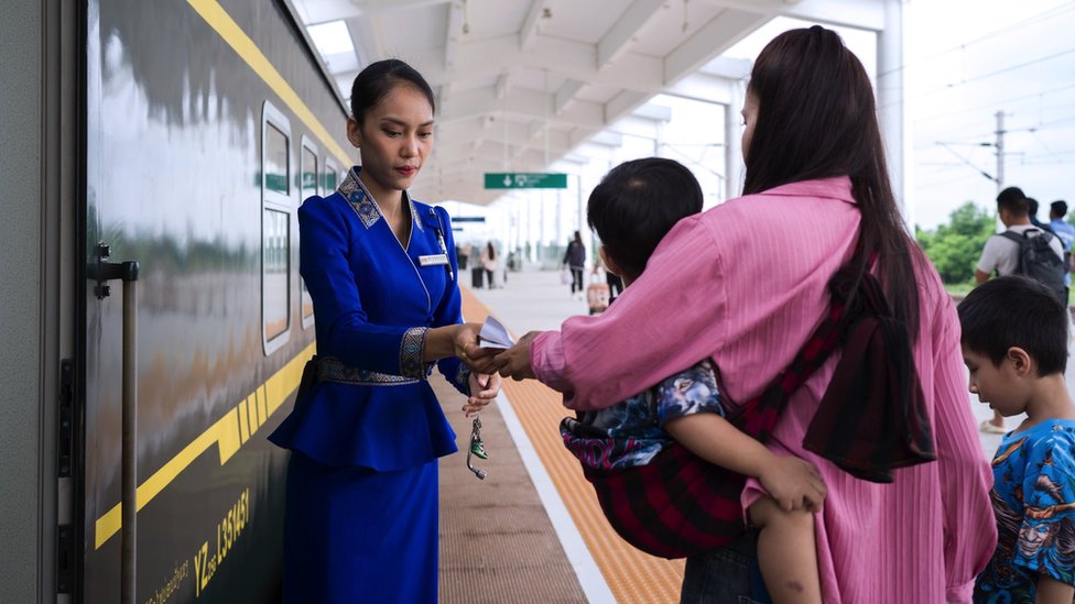 A woman and two young boys with her waiting to board the train at the Vientiane high speed railway station, the woman handing her ticket to a female inspector in uniform