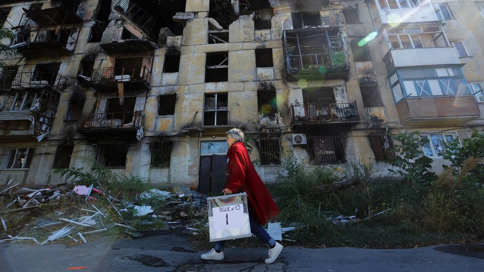 在烏克蘭馬里烏波爾，一名婦女提著便攜選票袋經過一座嚴重受損的居民樓