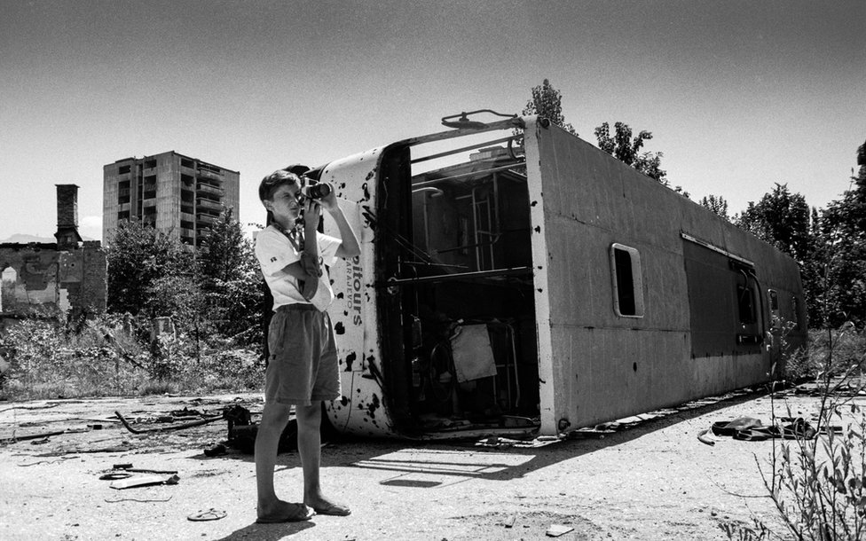 Ребенок держит фотоаппарат рядом с разрушенным автобусом