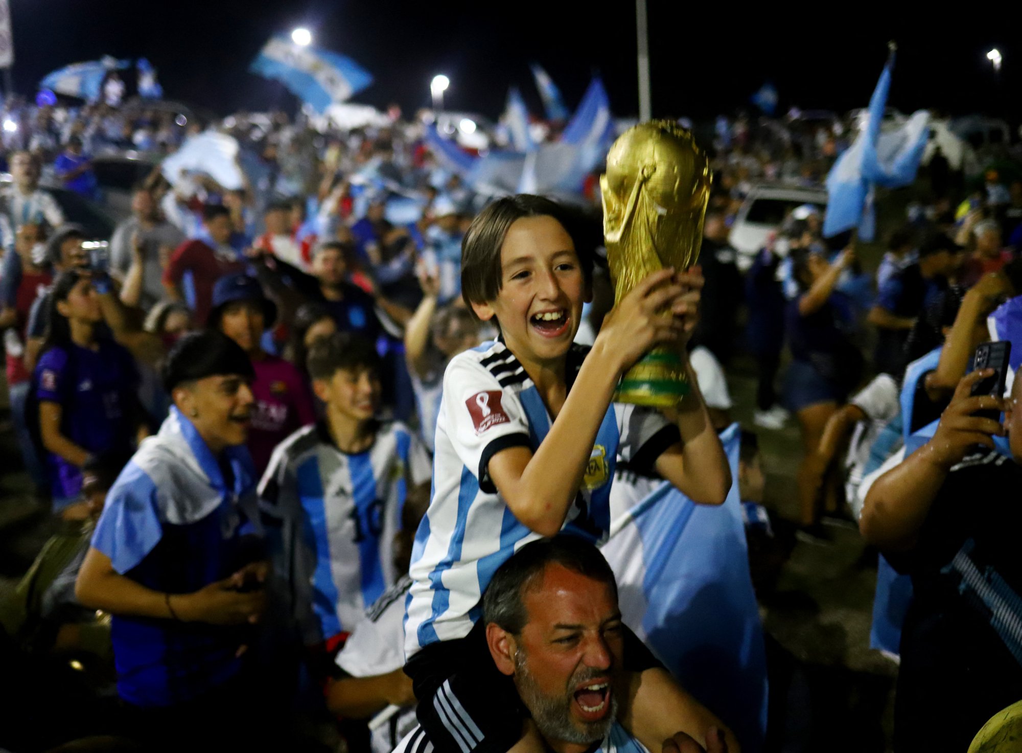 Un joven sobre los hombros de un adulto sostiene una réplica de la Copa FIFA