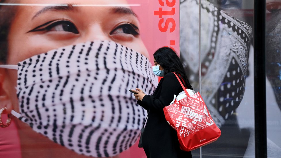 Woman in a mask walking past a store with an advertisement asking people to stay safe and wear a mask