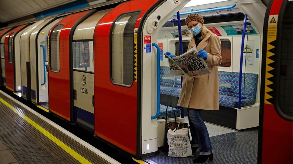 Женщина в СИЗ (средствах индивидуальной защиты), в том числе в маске в качестве меры предосторожности против COVID-19, читает газету, стоя в поезде лондонского метро, ??i