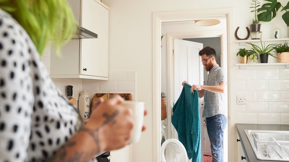 Mulher na cozinha tomando café enquanto homem lava a roupa